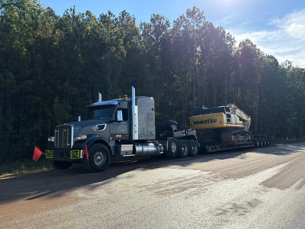 Silver truck hauling a Komatsu excavator on a lowboy trailer beside a wooded road