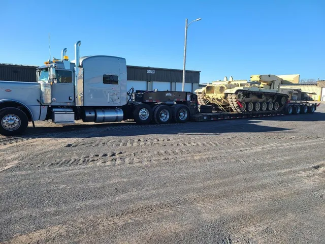 Silver truck hauling a tracked military vehicle on a lowboy trailer