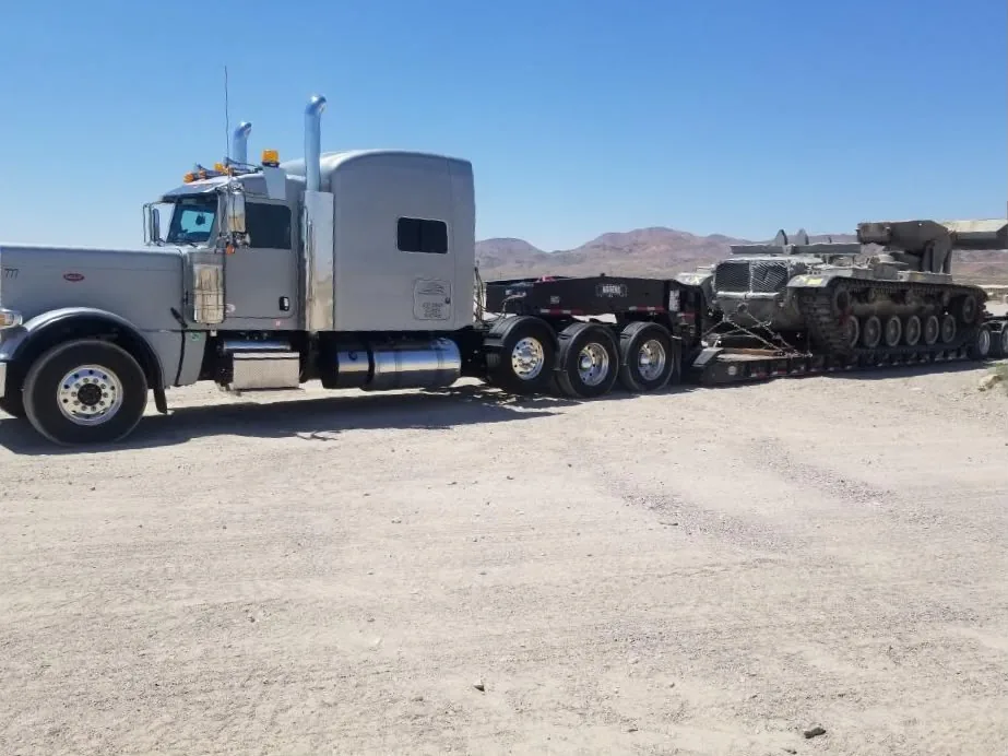 Side view of a silver truck and tracked military vehicle on a trailer